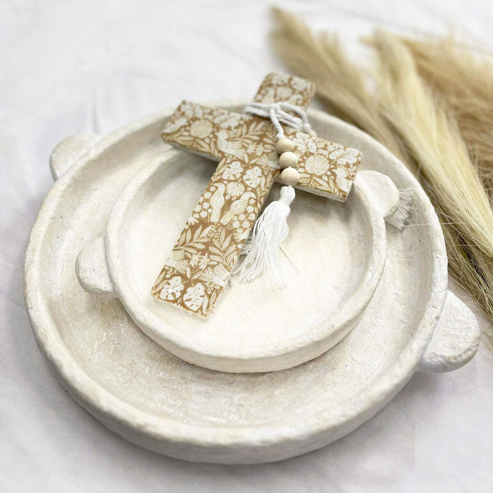 A decorative wooden cross ornament with a beige and white floral pattern, a white tassel, and a small wooden figure, displayed on a stack of rustic ceramic dishes with dried pampas grass in the background.