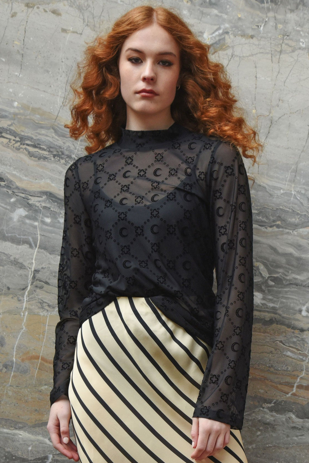 Woman wearing a black mesh flocked top and striped skirt against a stone wall.