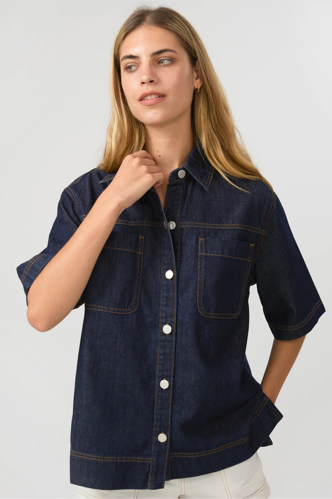 A woman wearing a dark indigo denim short-sleeve button-up shirt with contrast stitching and white buttons, paired with off-white pants, against a light gray studio background.