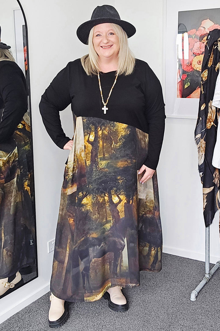 Woman wearing a black top and patterned skirt standing in a room with a mirror and clothing rack.