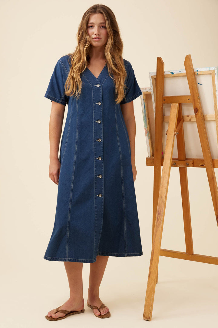 A woman modeling a short-sleeve denim midi dress with a button-down front, standing next to a wooden easel on a warm beige background.