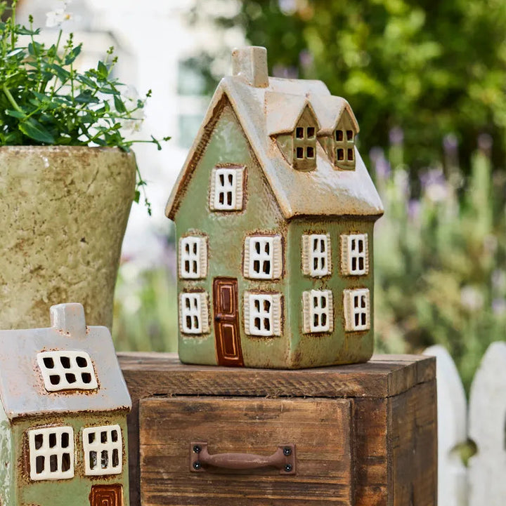 A decorative ceramic house-shaped figurine with a light green body, cream-colored textured trim, and a brown door, displayed on top of a rustic wooden chest outdoors.