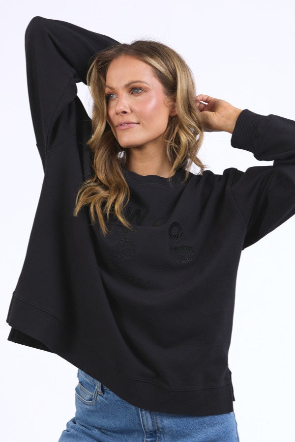 A female model wearing a black round neck crew neck jumper with chest embroidery, paired with blue jeans.