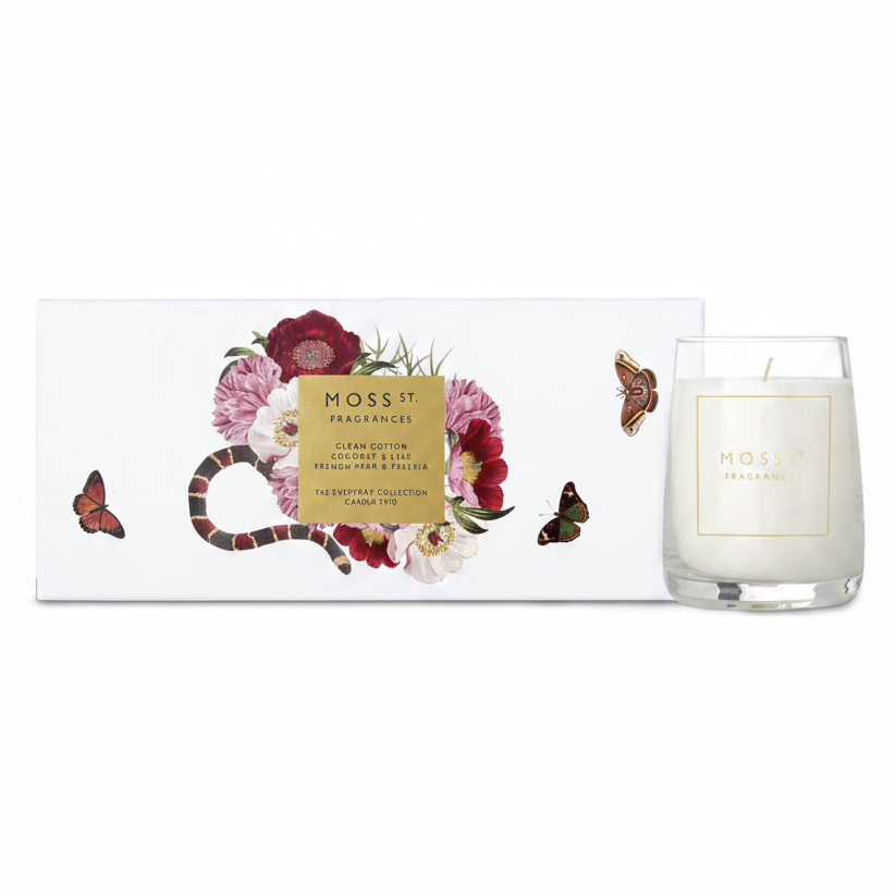 A white scented candle in a clear glass jar sits beside its white box packaging. The box features an illustration of a red and black banded snake wrapped through pink and burgundy flowers, with a central gold label reading 'MOSS ST.