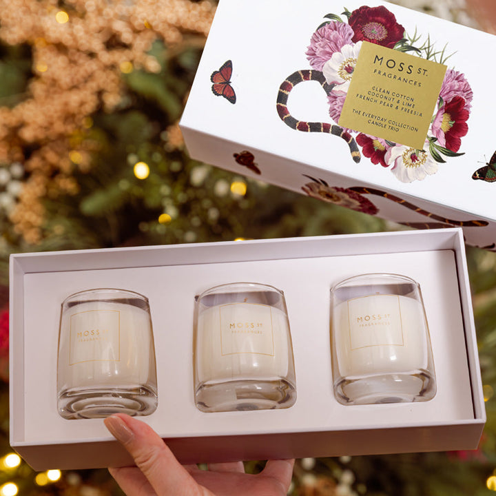 A hand holds an open gift box containing three small glass candles with cream wax. The box lid is placed above, showing floral artwork and a gold label that reads Mossy Fragrances.