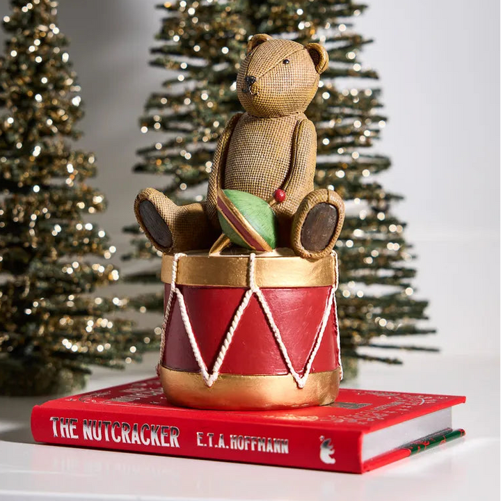 Decorative setup with a teddy bear on a drum, book titled 'The Nutcracker', and Christmas trees in the background.