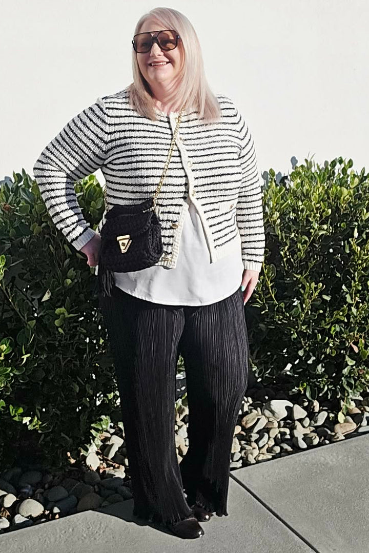 A woman posing outdoors on a concrete walkway wearing a black and white horizontally striped knit cardigan over a white top, black pleated trousers, and carrying a black crossbody bag.