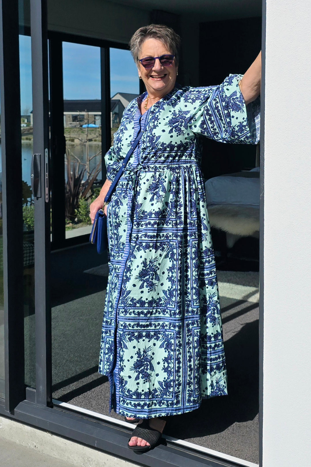 A woman wearing a blue and mint green patterned maxi dress with wide sleeves, standing in the doorway of a modern home.