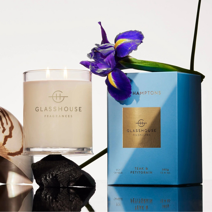 Glasshouse candles and a blue container with a purple flower on a reflective surface.