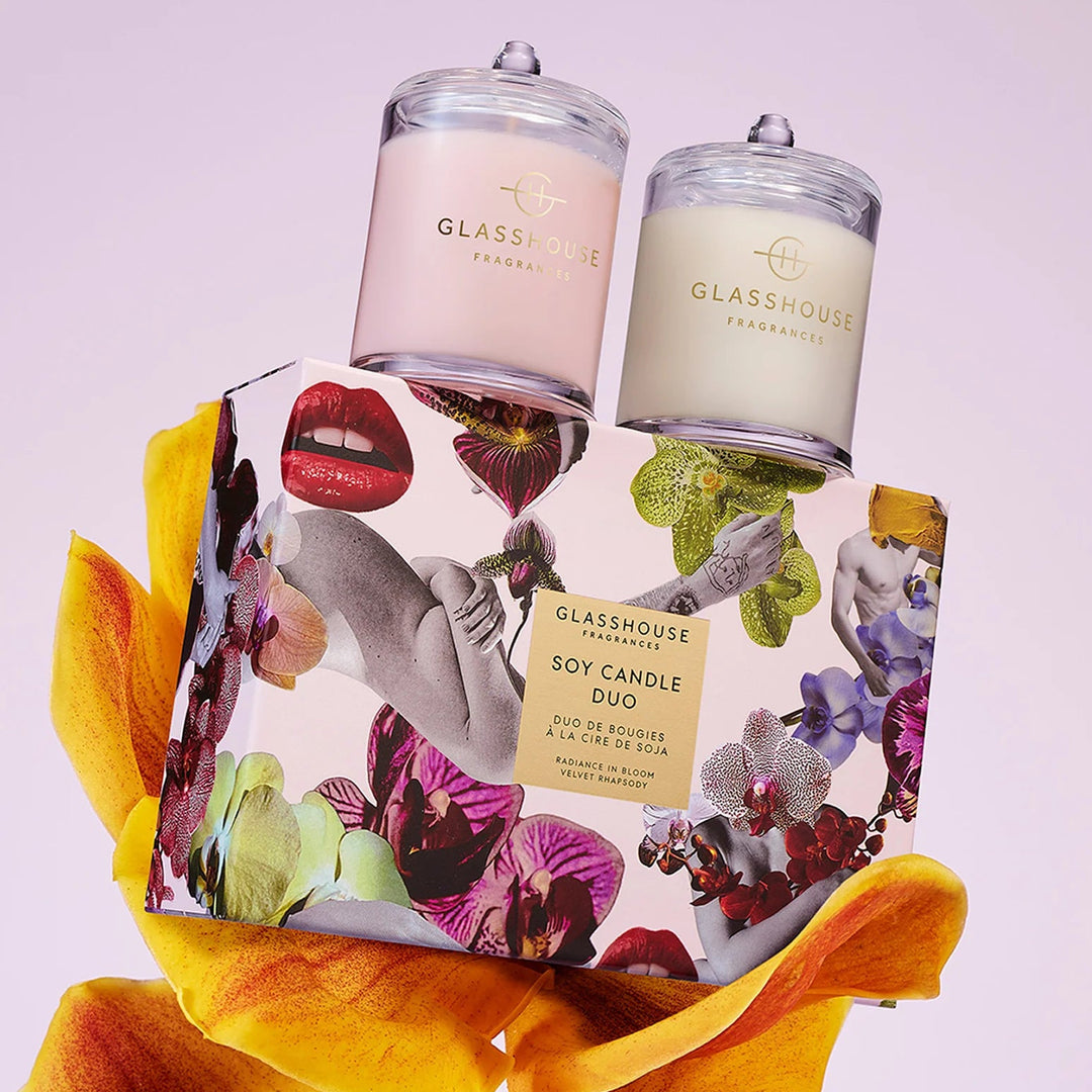 Two glass candles with floral packaging and a large yellow orchid on a light purple background.