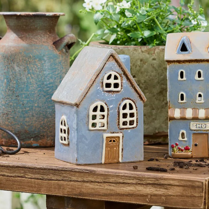 Two decorative miniature house figurines in light blue and beige tones, styled on a rustic wooden table with a blue ceramic vase and green foliage in the background.