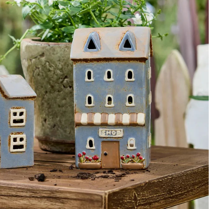 Two decorative miniature house-shaped garden planters sit on a rustic wooden table outdoors, with a blurred green garden and white flowers in the background.