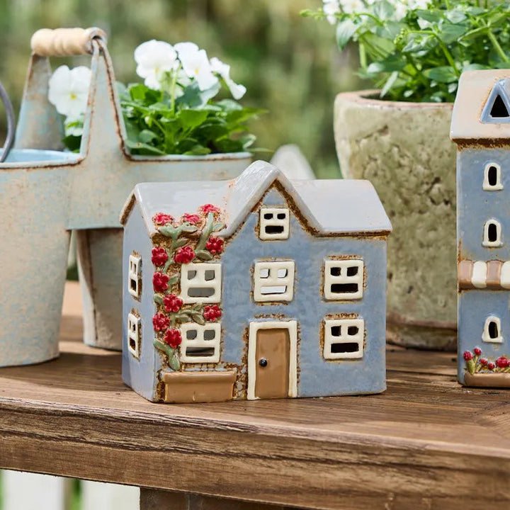 A decorative miniature house figurine in light blue and beige with red floral accents, displayed on a wooden table alongside other garden-themed ceramic decor and potted plants.