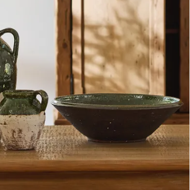 Ceramic vases and bowls on a wooden table with a plant and wooden cabinet in the background.