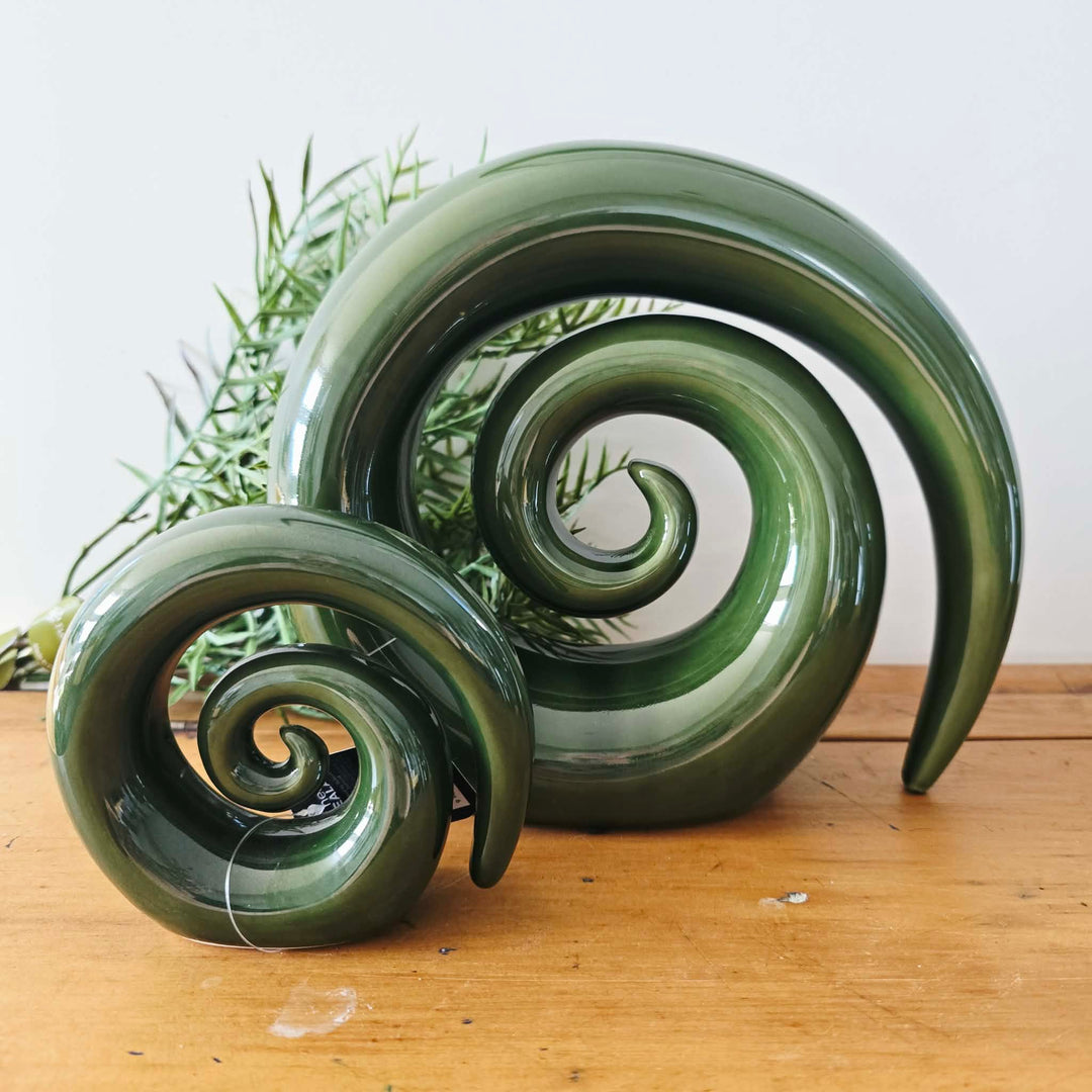 Two glossy dark green spiral decorative sculptures displayed on a wooden surface against a plain light background, with a small green plant behind them.