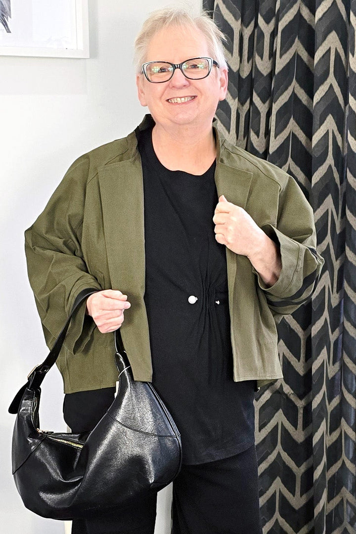 Person wearing a green jacket and black pants holding a black handbag in a room with patterned curtains.