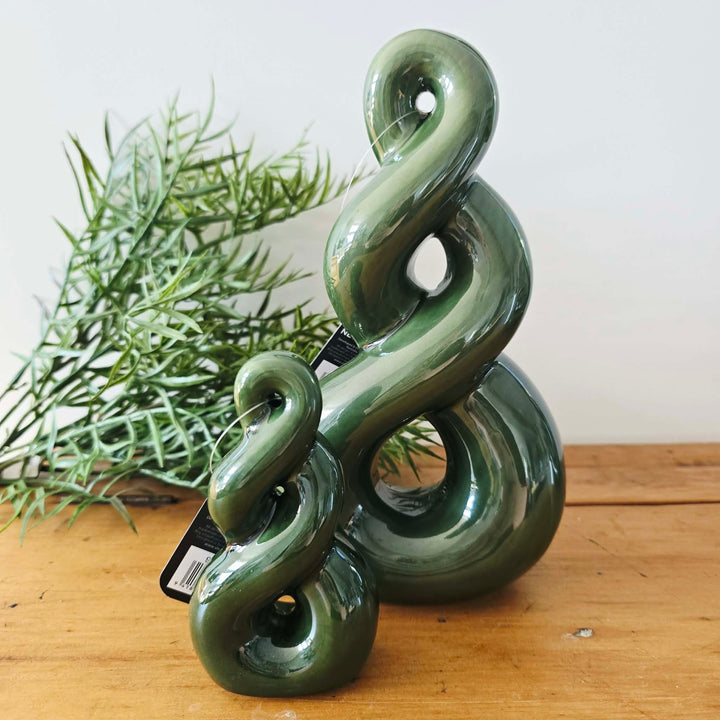 A glossy, sculptural green decorative object with an abstract, intertwined spiral shape, displayed on a wooden surface next to a green plant against a plain white wall.