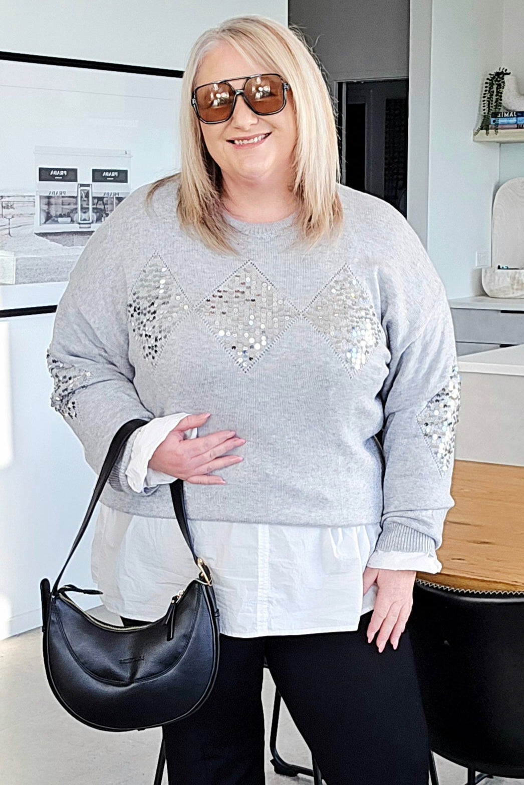 Woman in a gray sweater with embellishments, black pants, and sunglasses standing in a modern kitchen.