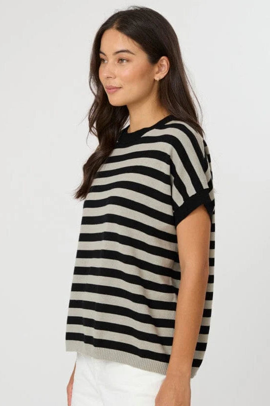 A woman wearing a short-sleeve knit top with horizontal black and beige stripes, paired with white pants, photographed against a plain light gray studio background.