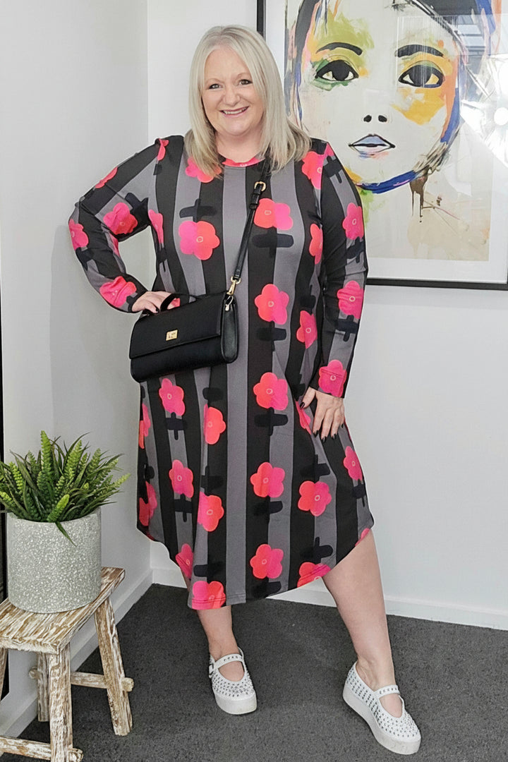 Woman wearing a black dress with pink floral patterns in a room with a plant and artwork on the wall.