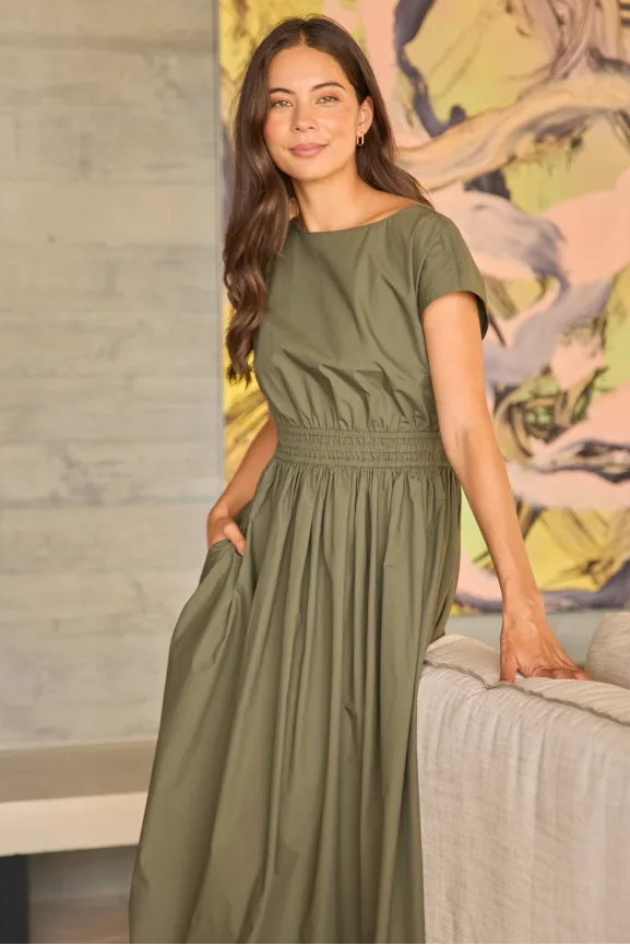 A woman wearing an olive green short-sleeve maxi dress with a smocked waistband, standing in a modern interior setting with a gray concrete wall and a colorful abstract painting in the background.