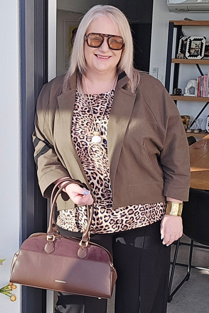 Woman holding a brown handbag, wearing a leopard print top and brown jacket, standing in front of a door.