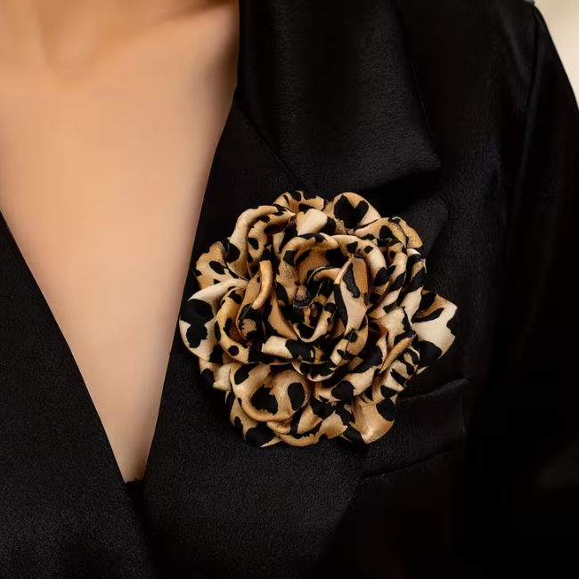 A close-up of a black satin lapel featuring a large fabric rose brooch with a beige and black leopard print pattern, worn on a model's chest.