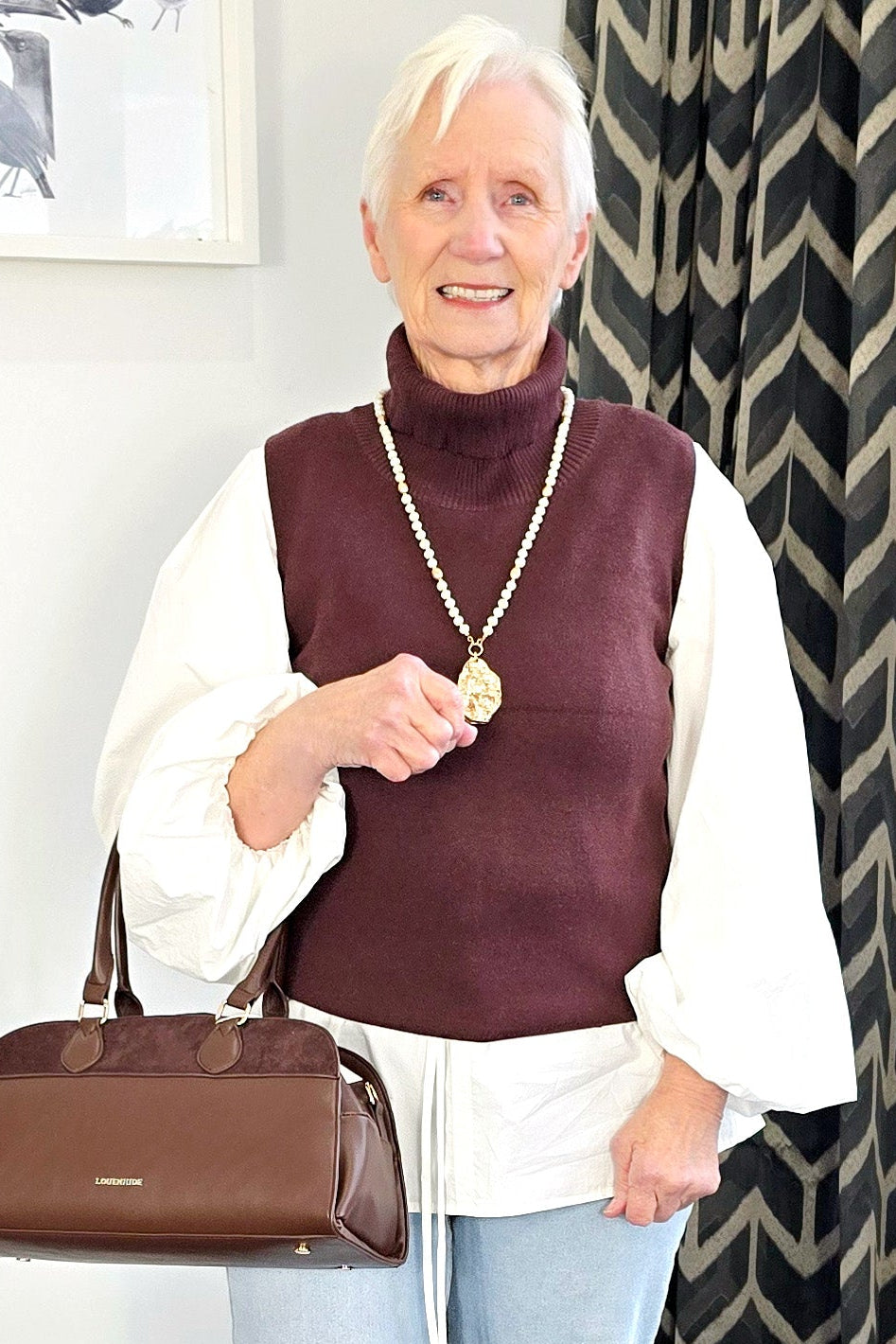 Woman holding a brown handbag indoors with a patterned curtain in the background