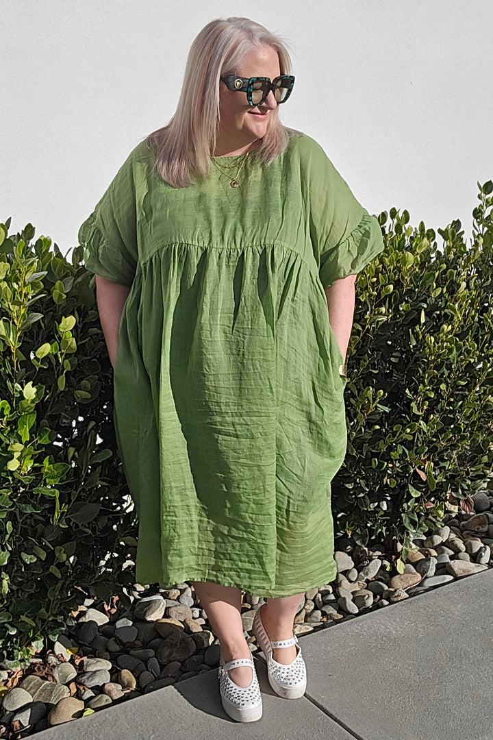 A woman with blonde hair and sunglasses wearing a loose, short-sleeved sage green dress, standing outdoors on a concrete path next to green bushes.