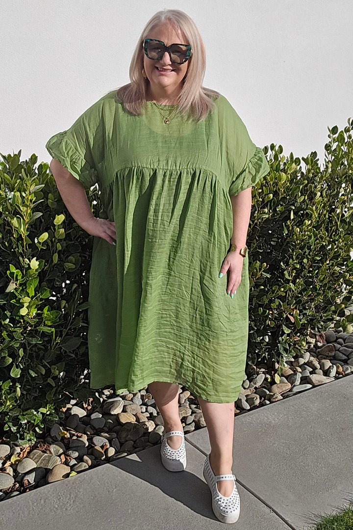 A woman wearing a loose, short-sleeve sage green midi dress and white sneakers stands outdoors on a concrete walkway next to green bushes against a plain white wall.