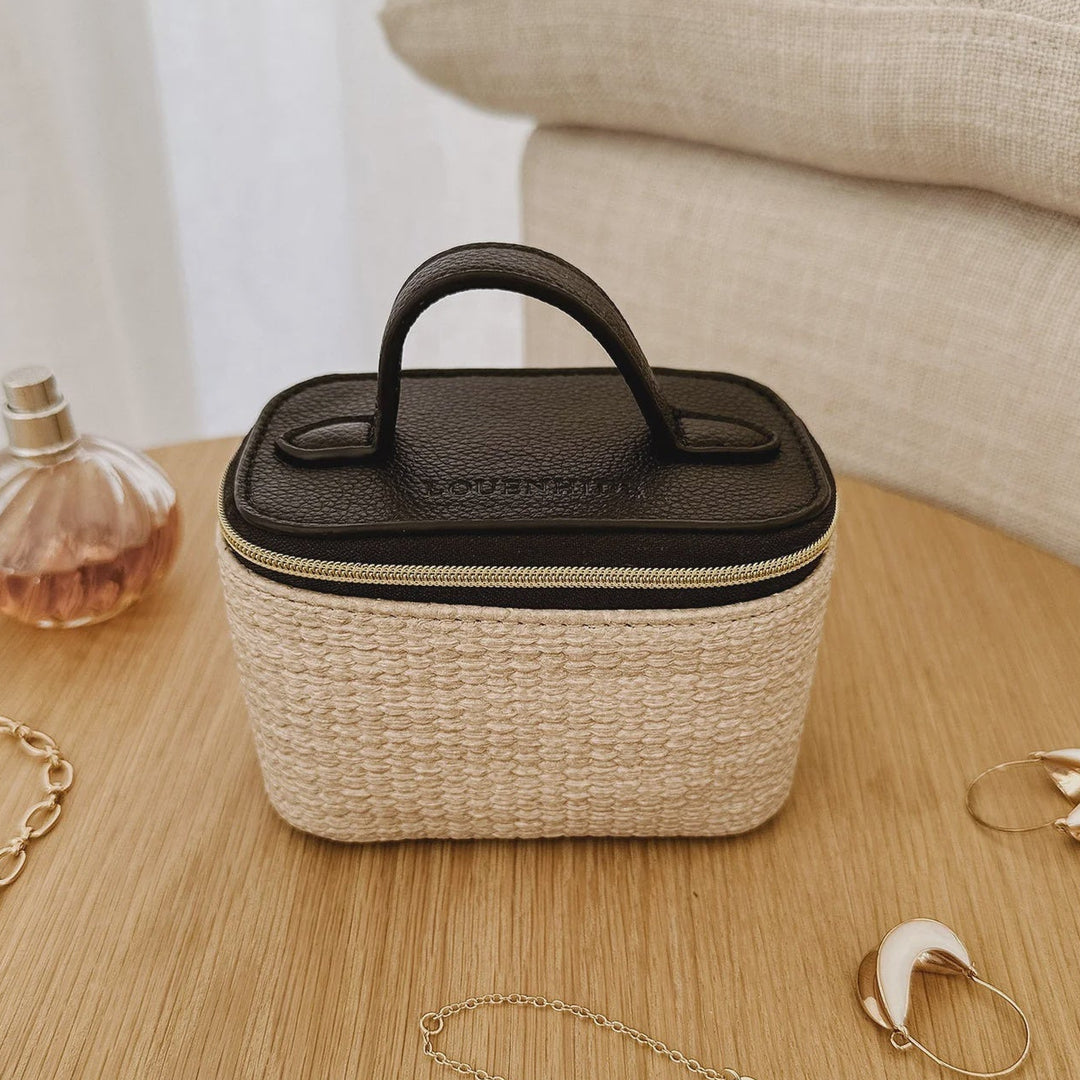 Small woven travel bag with black top on a wooden surface with jewelry and a perfume bottle.
