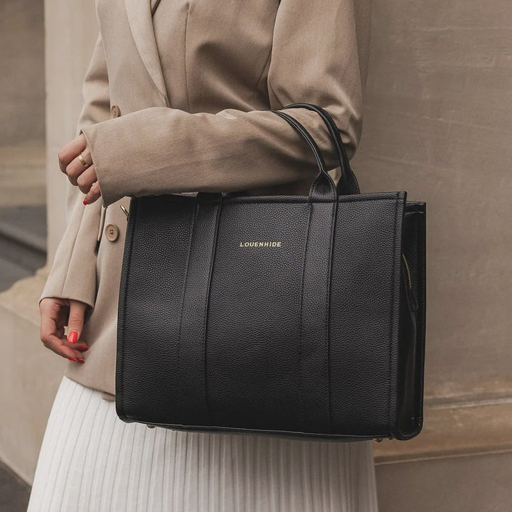 Woman holding a black louenhide tote bag with a brand logo, standing outdoors.