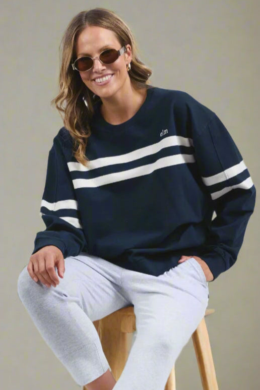 A woman sitting on a stool wearing a navy crew sweatshirt with white chest stripes and embroidery, paired with light-colored jeans and sunglasses.