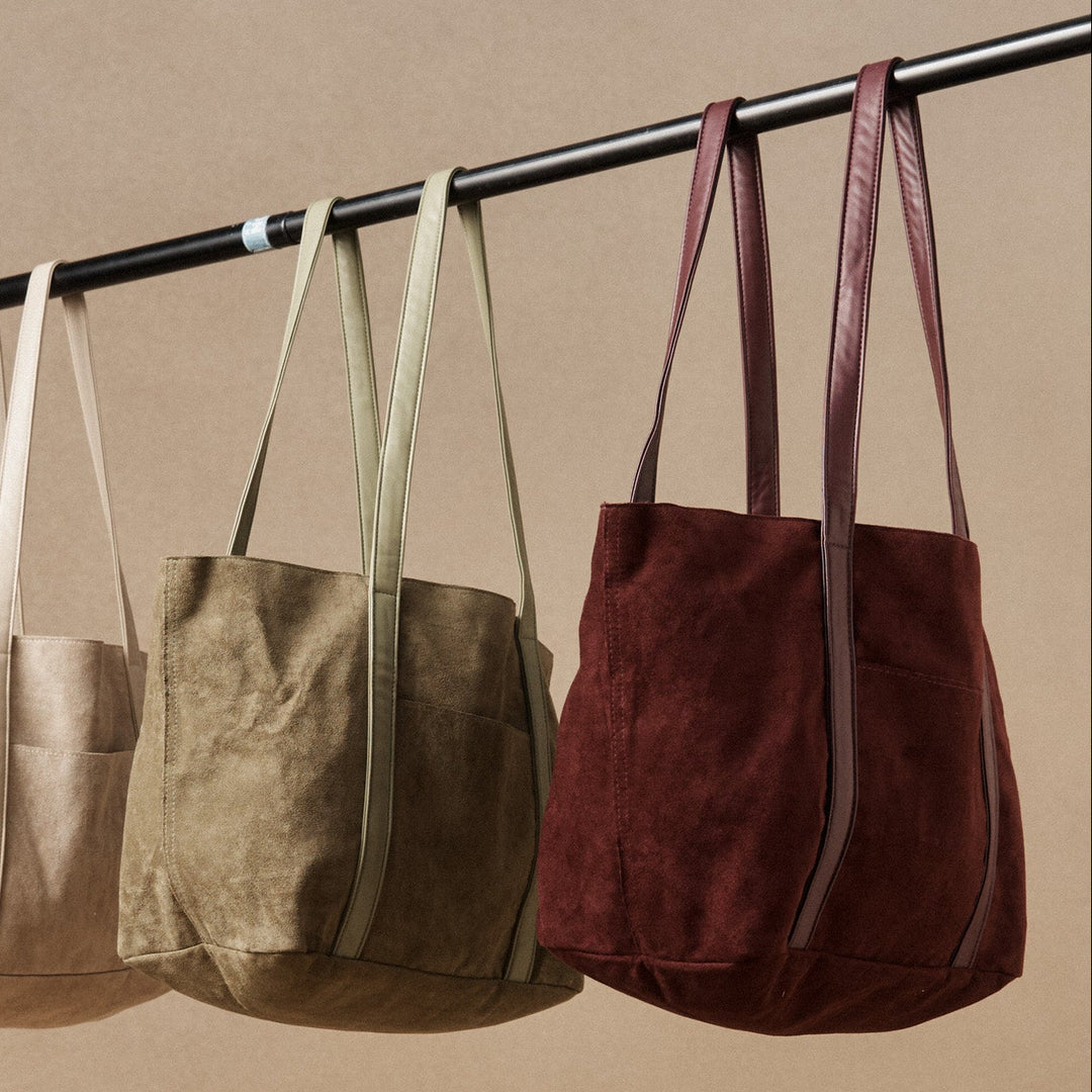 Three tote bags in beige, brown, and burgundy hanging on a rod against a beige background.