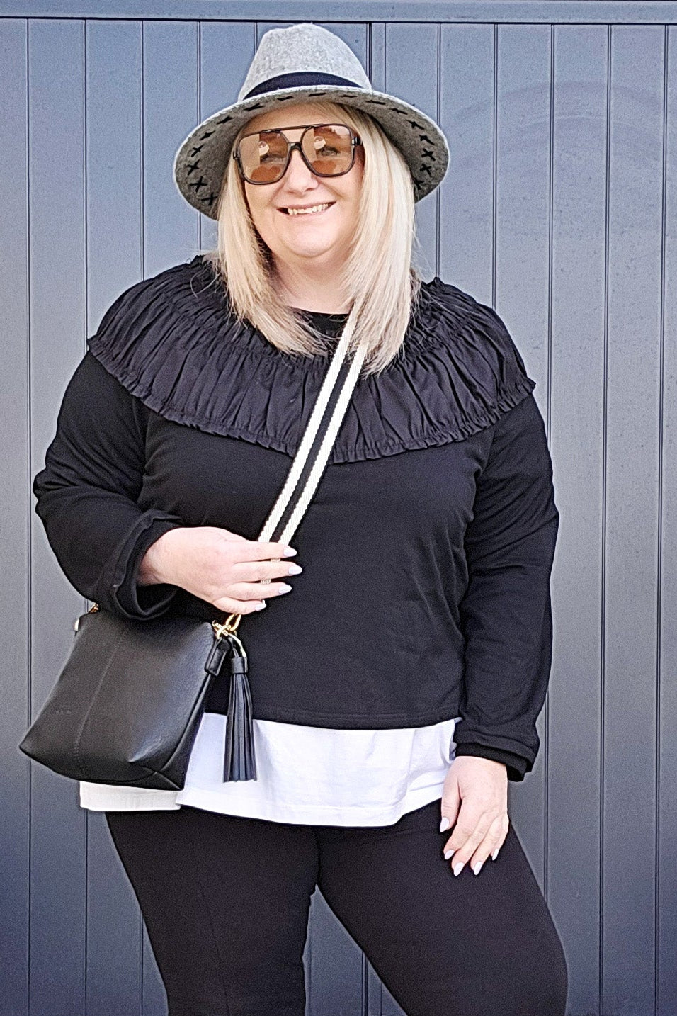 Woman in black outfit with white shirt, hat, and shoes standing against a blue wall.