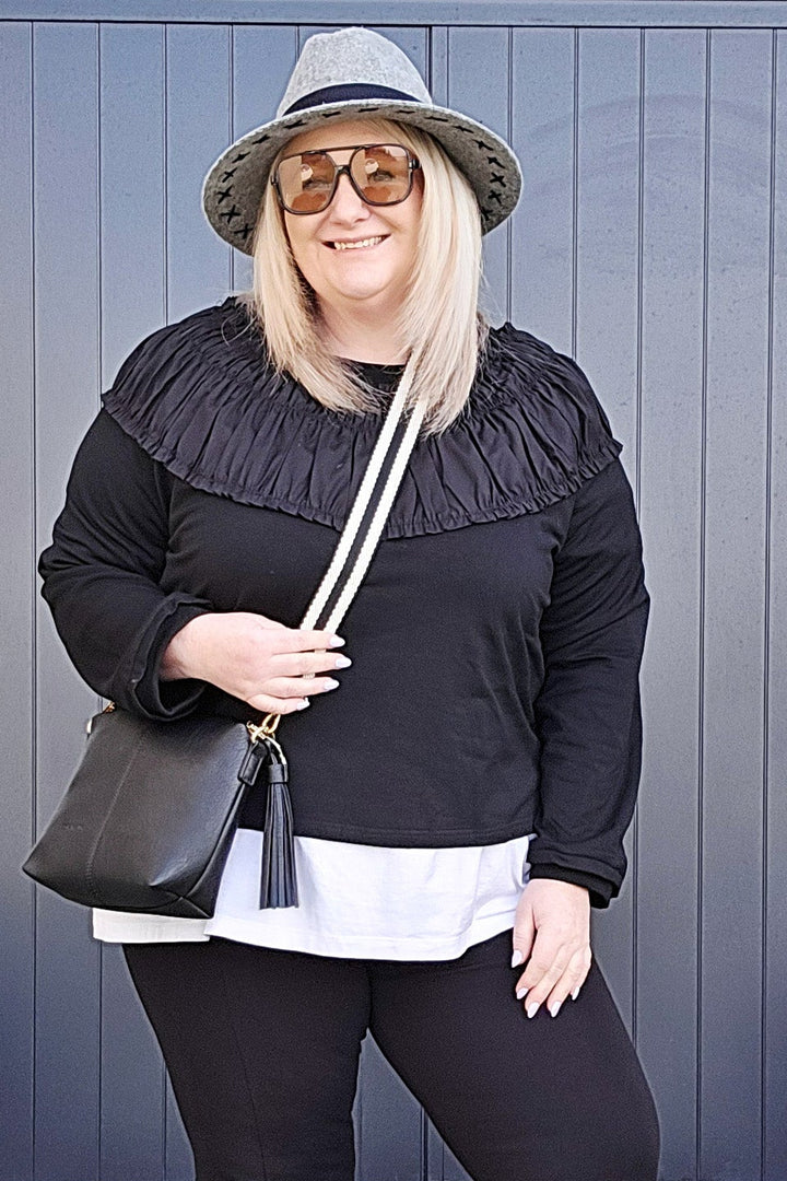 Woman in black outfit with white shirt, hat, and shoes standing against a blue wall.