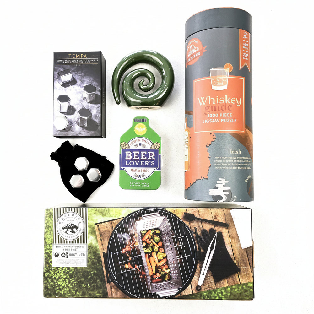A curated whiskey-themed gift set arranged on a light neutral background, including a cylindrical tin labeled 'Whiskey Quiz 2000 Piece Jigsaw Puzzle', a green spiral-shaped ceramic object, a black box labeled 'Tempa', a small black pouch with silver dice, a green and blue 'Beer Lovers' bottle-shaped item, and a rectangular box featuring a grill scene.