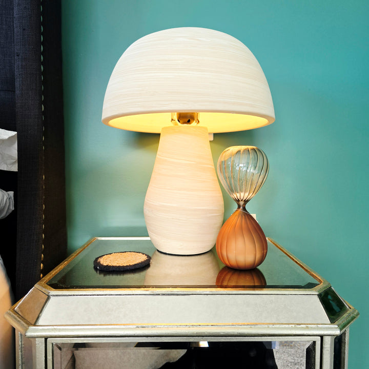 A table lamp with a textured cream-colored shade and base, glowing with a warm yellow light, displayed on a mirrored side table next to an hourglass against a teal wall.