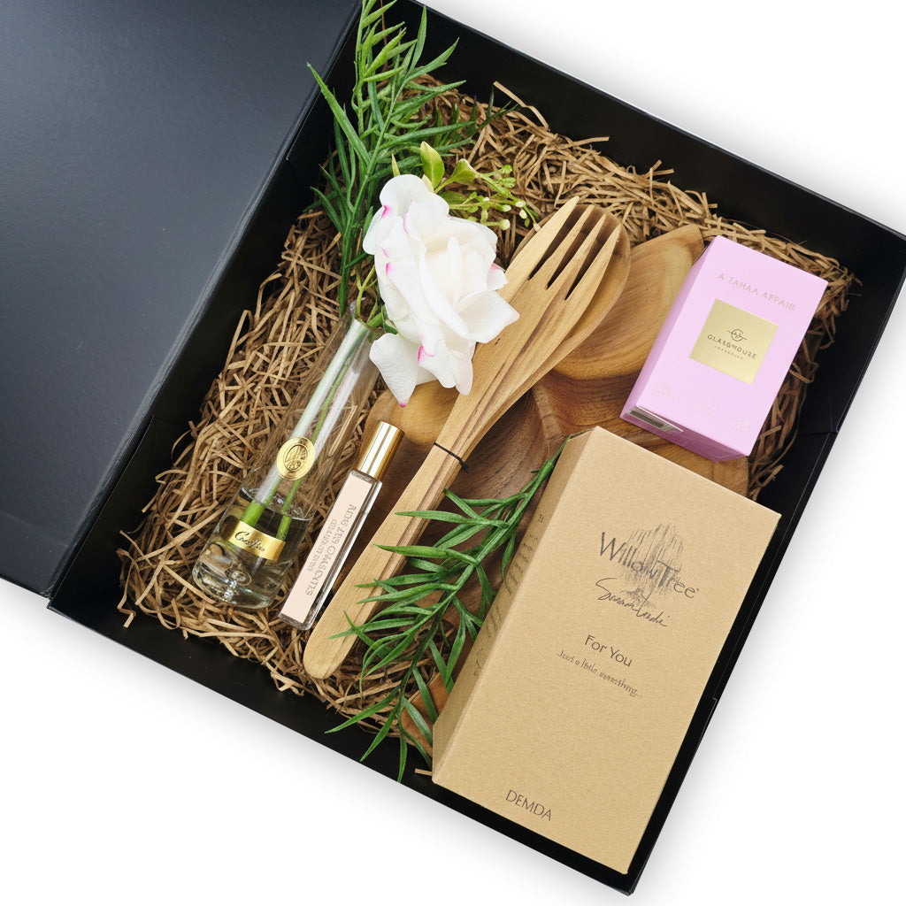 A curated gift set in a black box with shredded paper filler, featuring a small clear perfume bottle with a gold cap, a pink boxed item, wooden utensils, a kraft card, green sprigs, and a white flower on a white background.
