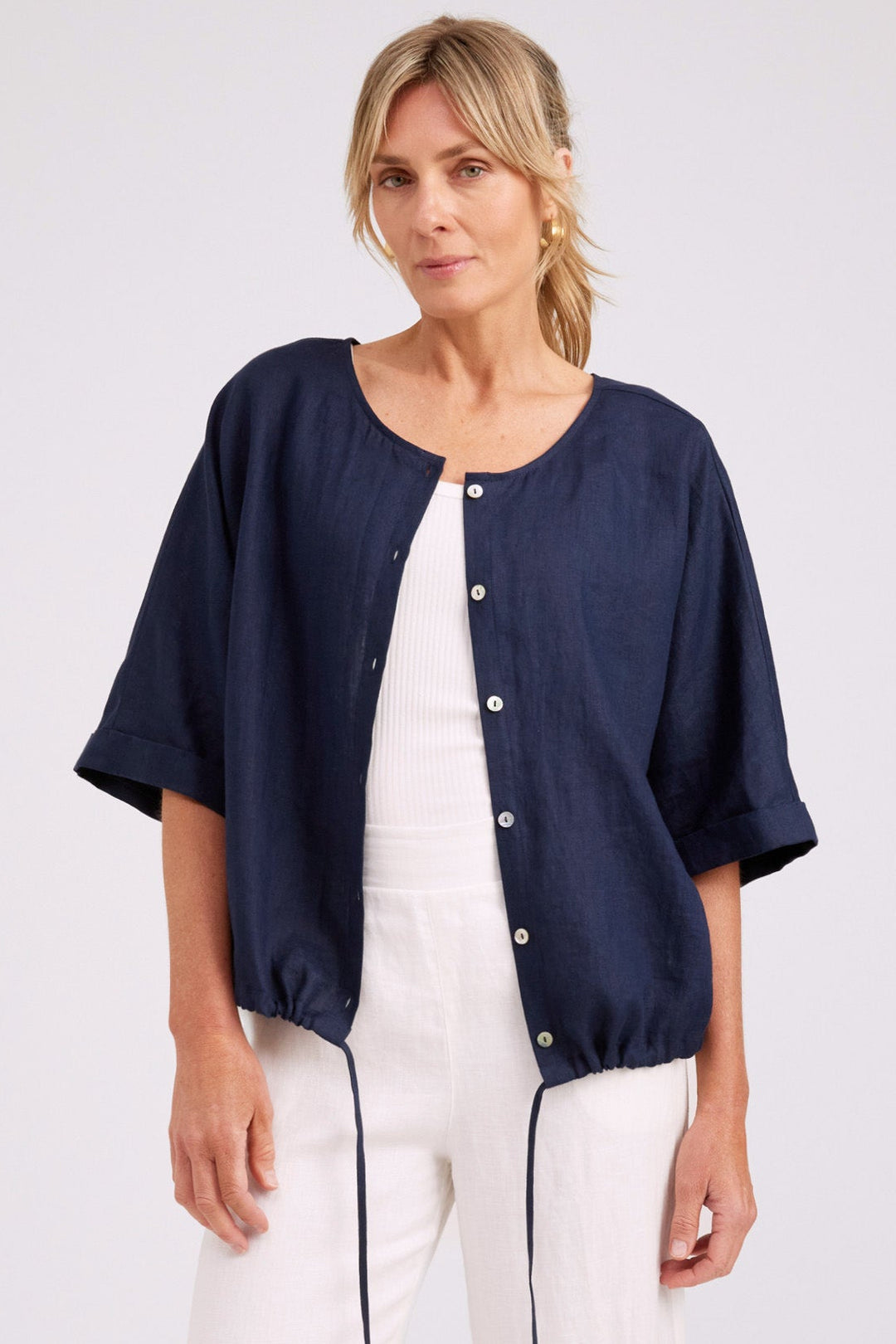 A woman modeling a navy blue short-sleeve button-front jacket over a white jumpsuit, photographed against a plain light gray studio background.