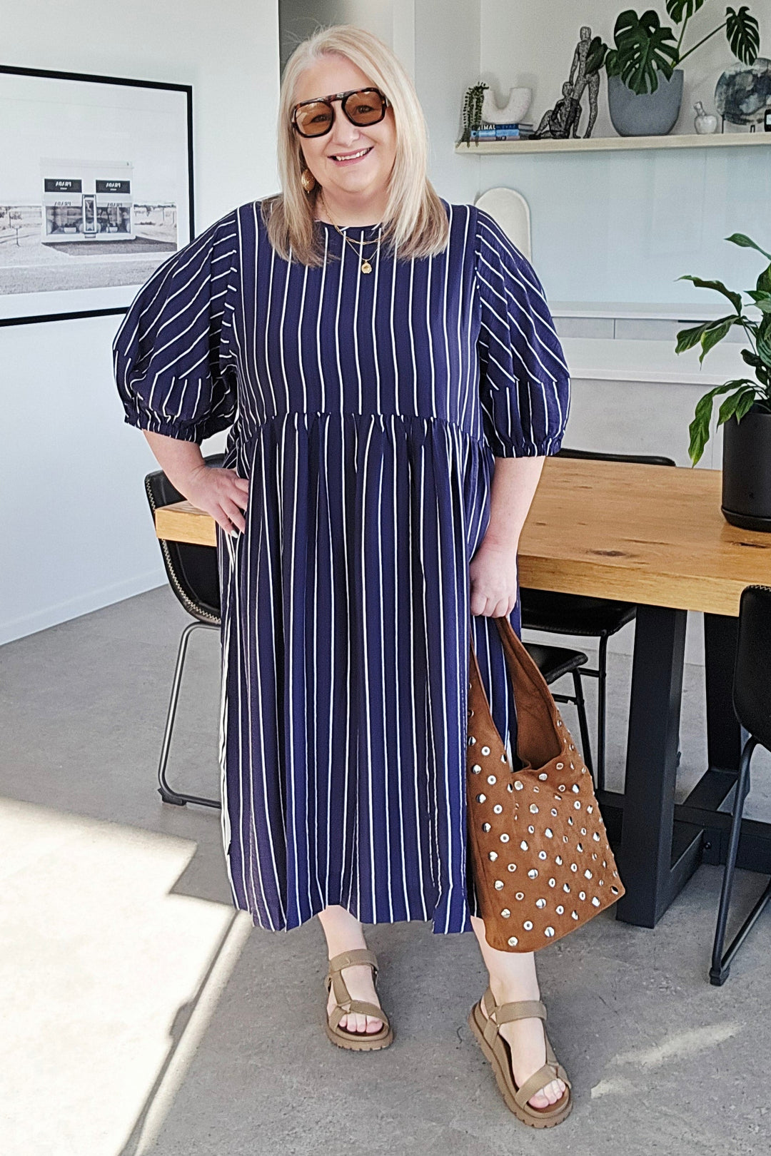 A woman wearing a navy blue and white vertically striped midi dress with puff sleeves, standing in a modern kitchen and dining area while holding a brown polka-dot tote bag.