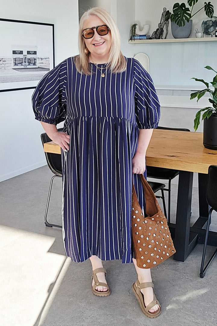A woman wearing a navy blue and white vertically striped midi dress with puff sleeves, standing in a modern kitchen and dining area while holding a brown polka-dot tote bag.