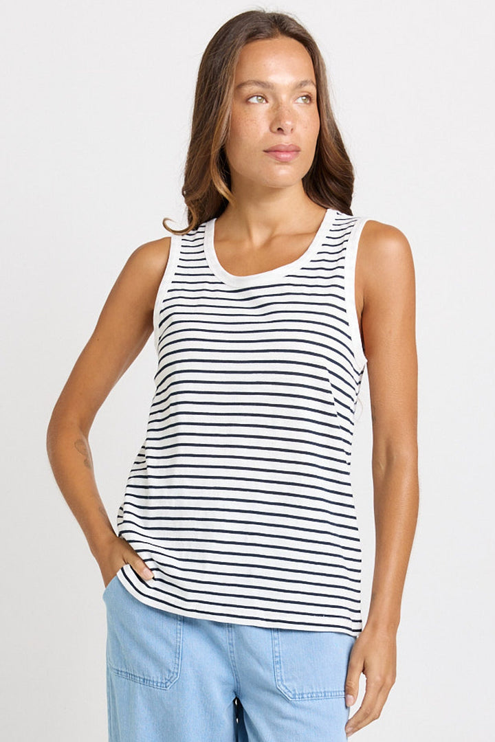 A woman modeling a sleeveless tank top with thin horizontal navy blue stripes on a white background, paired with light blue jeans, against a light gray studio backdrop.
