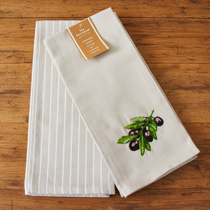 Two folded cotton kitchen towels by The Art of Living. One towel features a light beige and white vertical stripe pattern, while the other is a solid cream color with an embroidered olive branch motif with green leaves and dark purple olives.