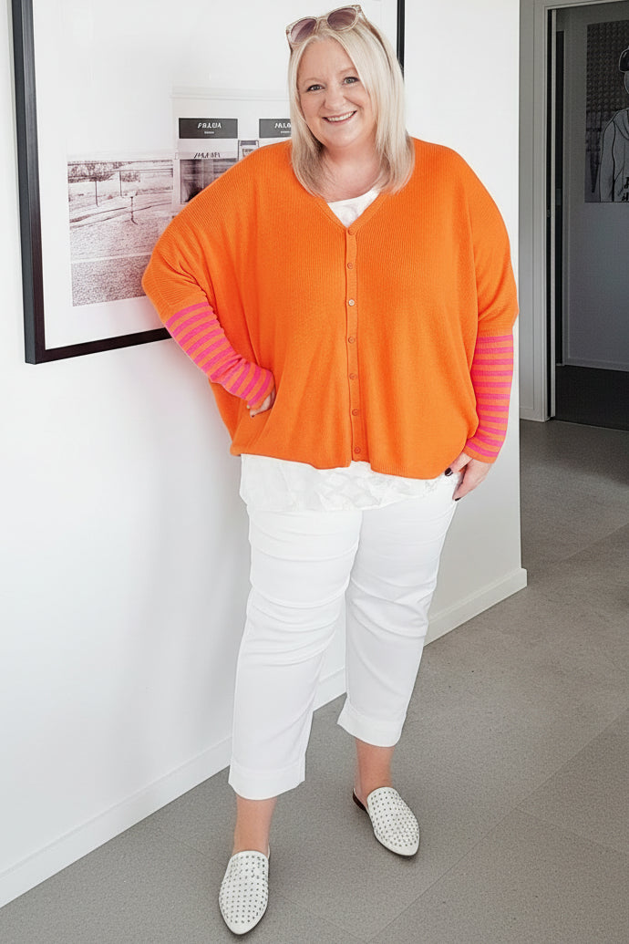 Woman wearing an orange cardigan with pink sleeves and white pants standing indoors.