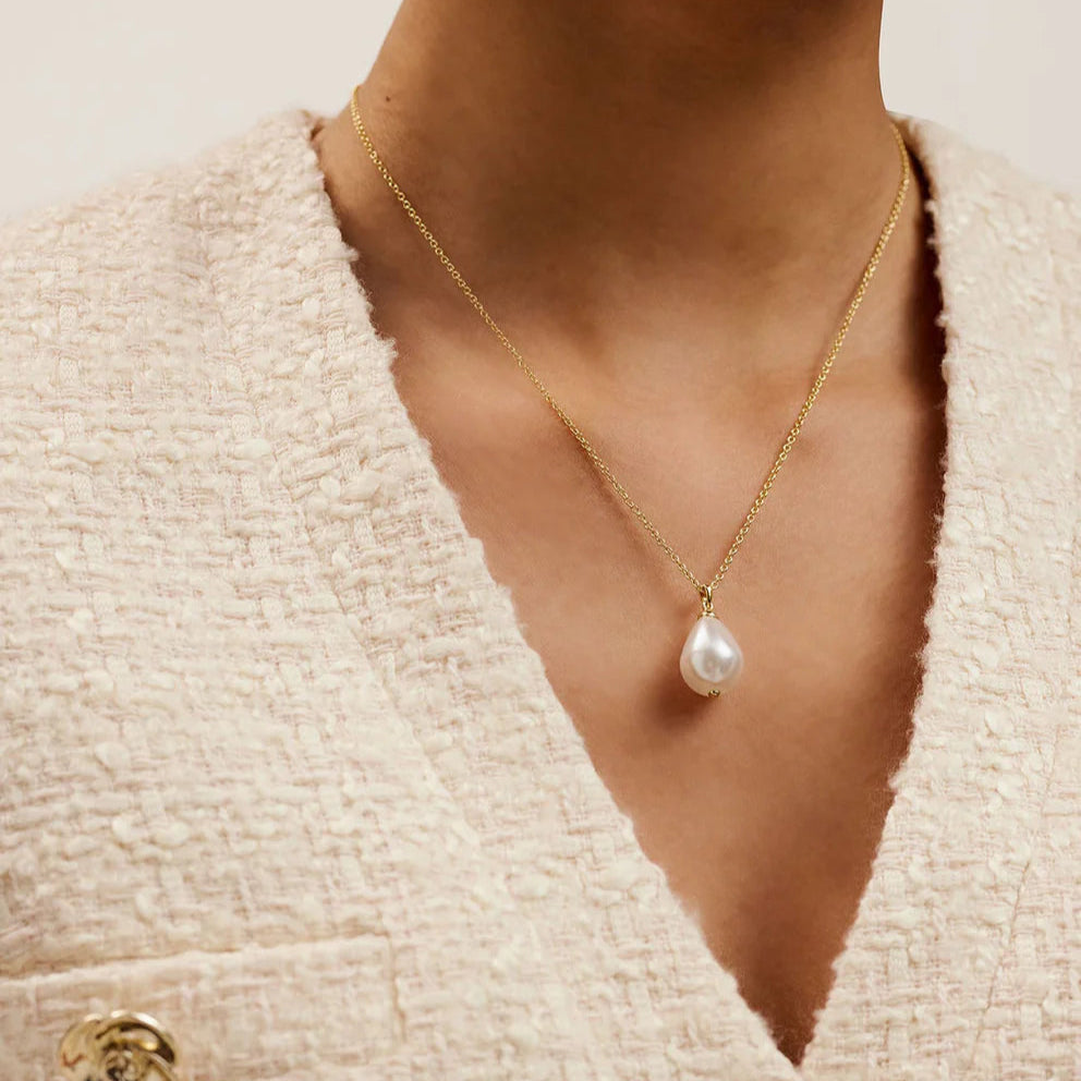 Gold necklace with a pearl pendant worn by a person in a textured beige cardigan.
