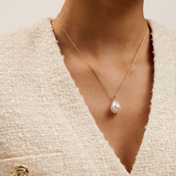 Gold necklace with a pearl pendant worn by a person in a textured beige cardigan.