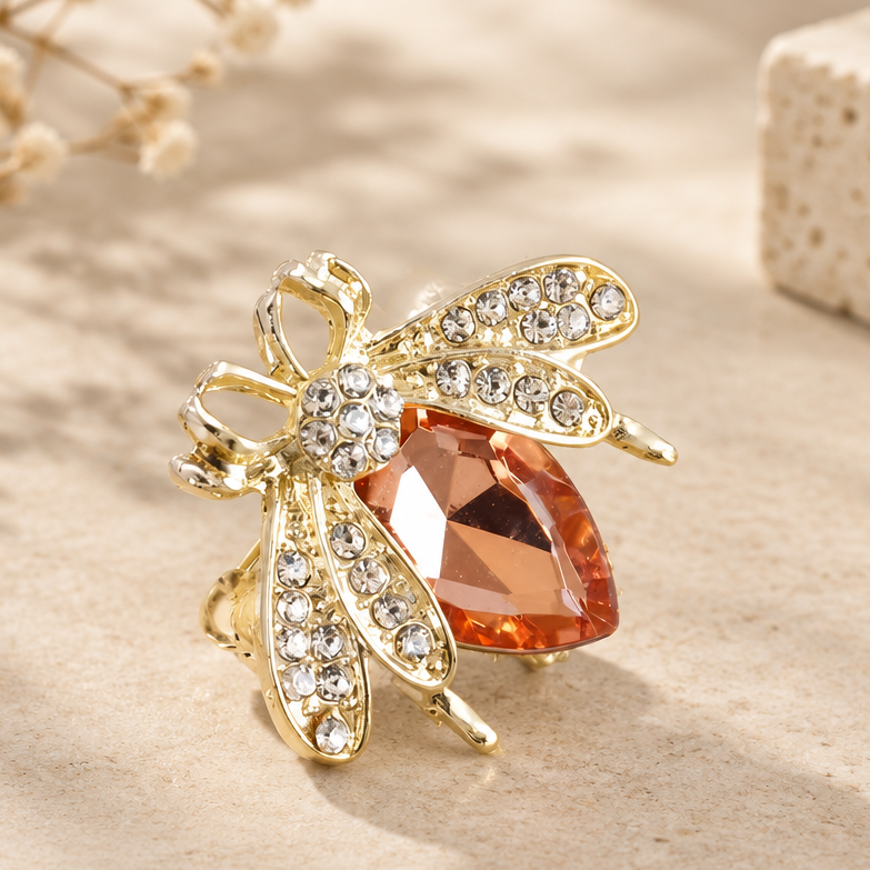Gold-tone bee-shaped ring with clear rhinestones and a peach-colored faceted crystal on a light gray textured background.