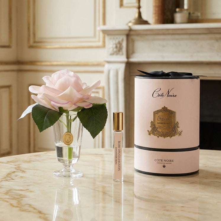 A gift set featuring a pale pink rose in a clear glass vase, a small perfume sample vial with a gold cap, and a tall cylindrical Côte Noire perfume box in pale pink with black ribbon and gold detailing, all on a white background.