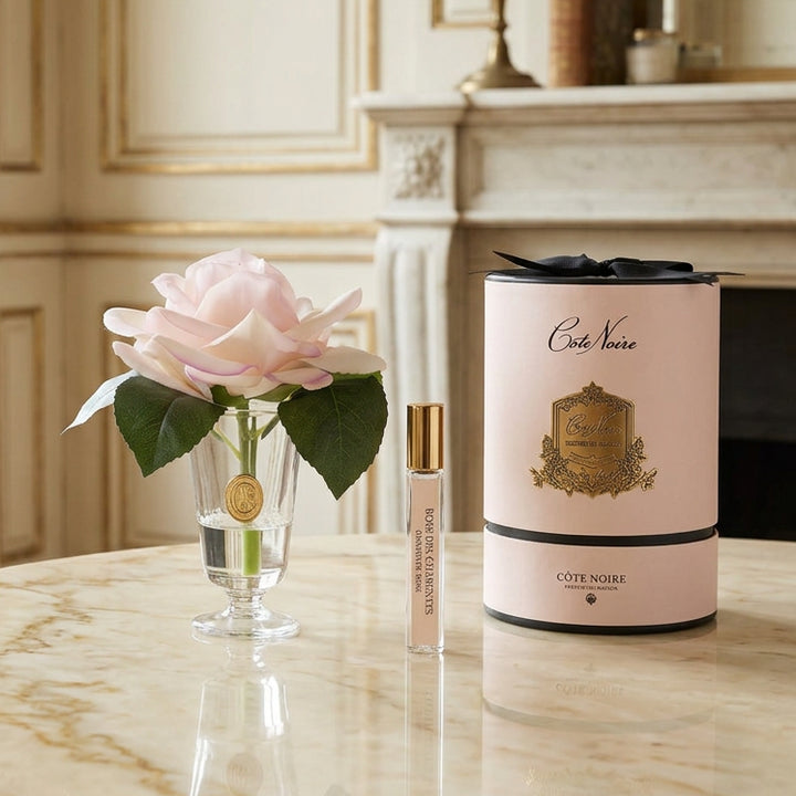 A gift set featuring a pale pink rose in a clear glass vase, a small perfume sample vial with a gold cap, and a tall cylindrical Côte Noire perfume box in pale pink with black ribbon and gold detailing, all on a white background.